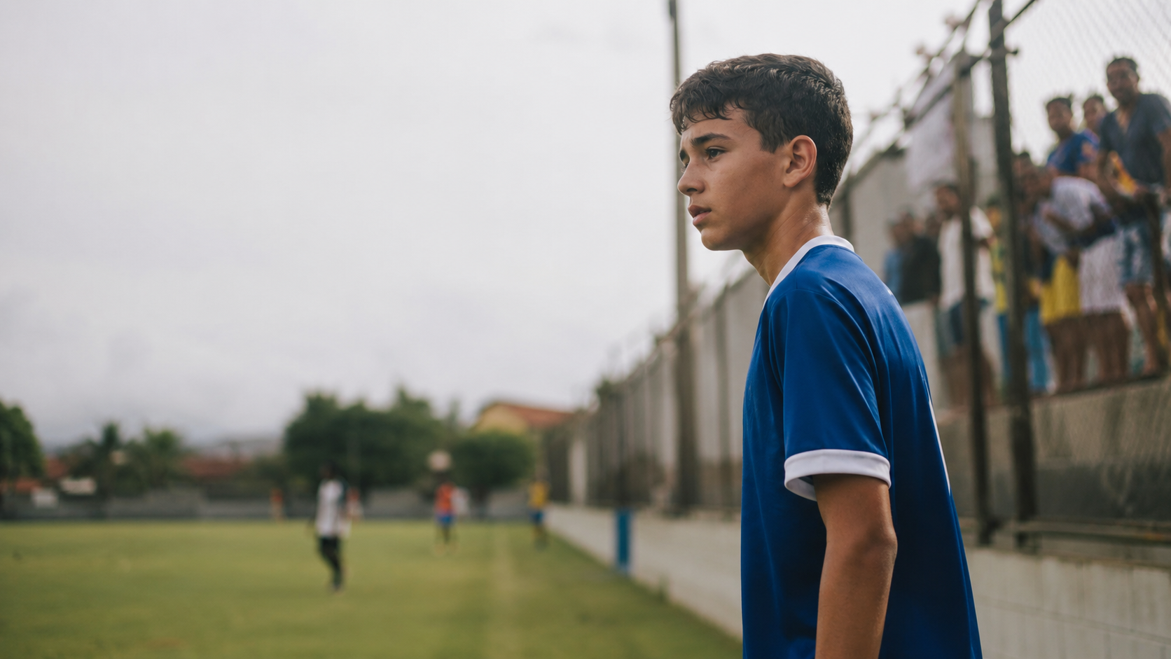 Jovem atleta de futebol olha para fora do campo enquanto adultos acompanham a partida ao fundo